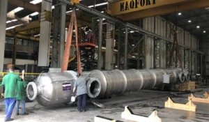 Trocador de calor industrial sendo manuseado em fábrica da Apema, com trabalhadores usando capacetes e macacões verdes, ilustrando um artigo sobre serviço de reforma em trocadores de calor.