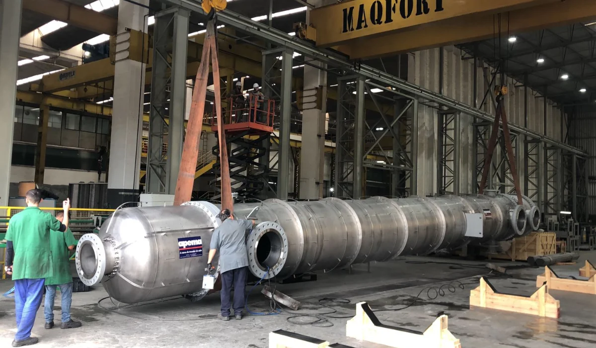 Trocador de calor industrial sendo manuseado em fábrica da Apema, com trabalhadores usando capacetes e macacões verdes, ilustrando um artigo sobre serviço de reforma em trocadores de calor.