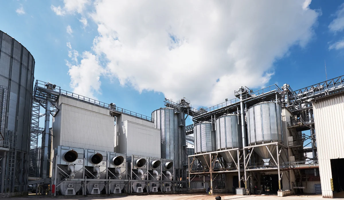 Indústria com silos metálicos e equipamentos de processamento sob céu azul com nuvens, que ilustram um conteúdo da Apema sobre usina de açúcar e álcool.
